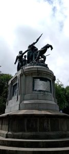 A bronze monument with statues on a stone pedestal in a public park.
