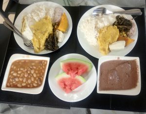 Two plates of a Ugandan traditional meal of rice, matooke, cassava, pumpkin, leafy greens, and groundnut sauce, served alongside beans and slices of watermelon.

