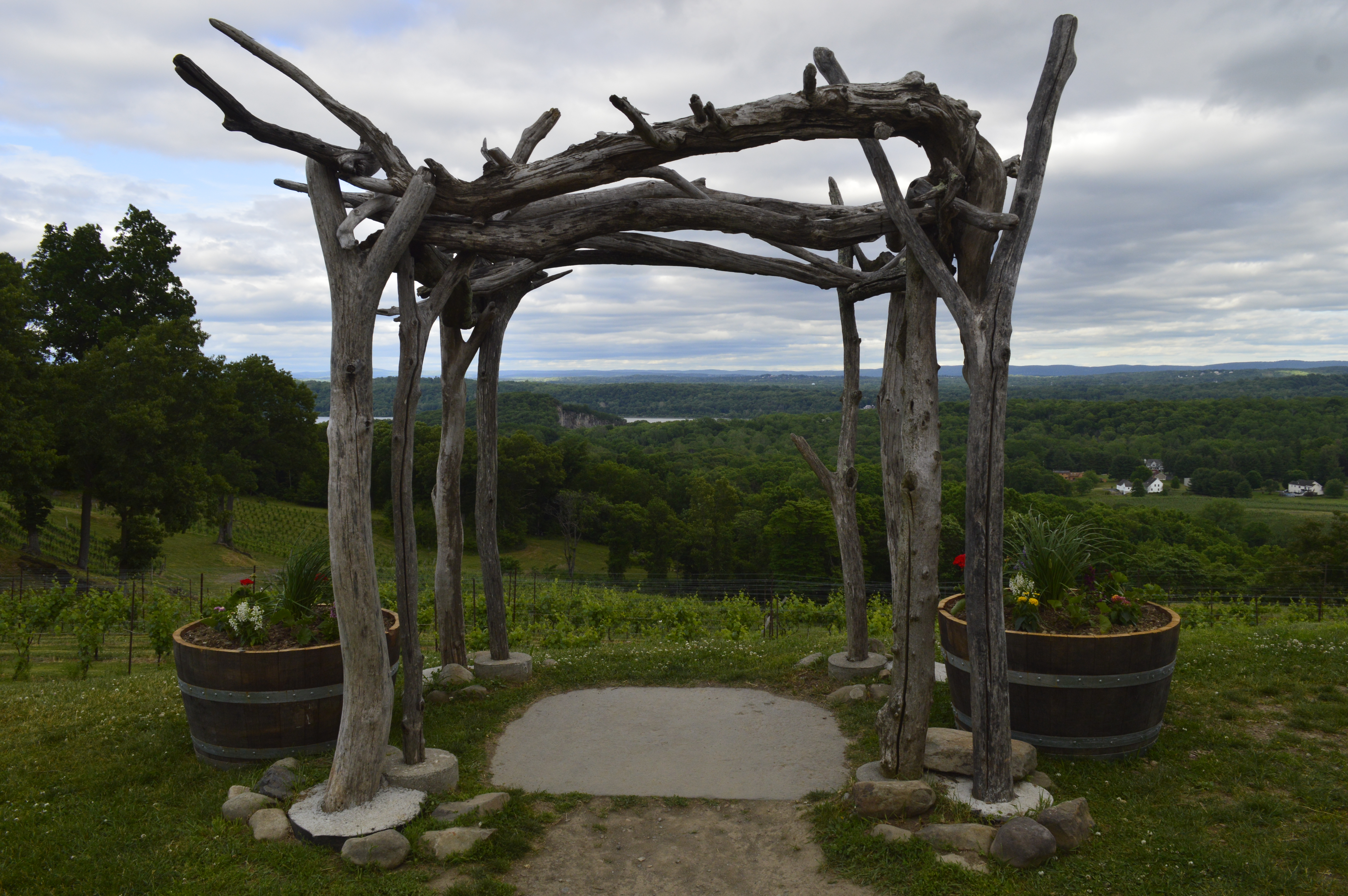 Rustic wooden arbor overlooking vineyards and rolling green countryside beneath a cloudy sky.