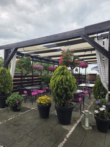 A covered rooftop terrace with flowers, outdoor chairs, and city rooftops in the background.
