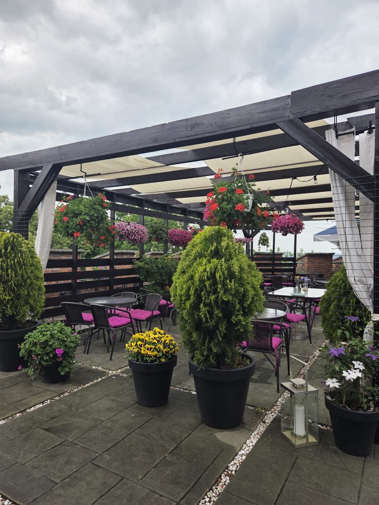 A covered rooftop terrace with flowers, outdoor chairs, and city rooftops in the background.
