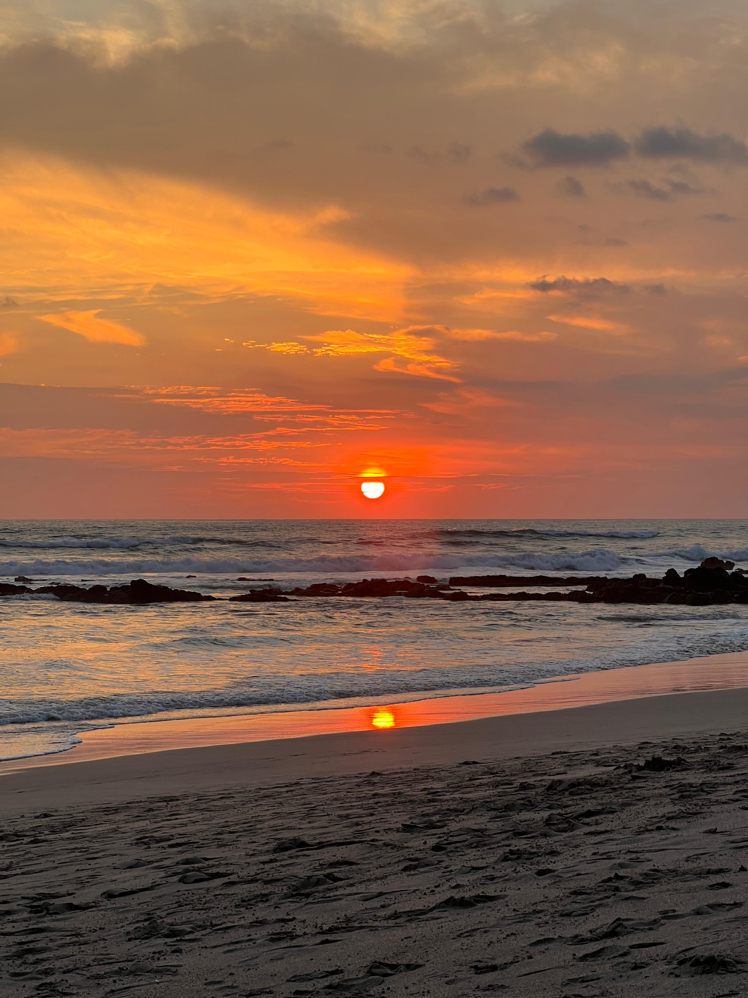 A serene beach scene at sunset, where the sun hangs low on the horizon, casting a warm, golden light that transitions into shades of orange, pink, and soft blue in the sky. The reflective surface of the water mirrors the colors of the sunset, creating a gentle shimmering effect along the shoreline. Waves softly lap against the sandy beach, leaving