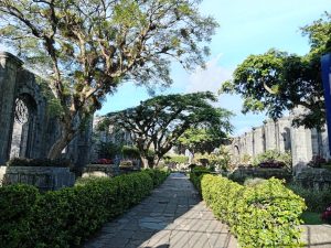 Stone pathway through the garden inside the Santiago Apostle Ruins in Cartago, Costa Rica. Trimmed hedges, colorful flowers and large trees line the path, flanked by ancient stone walls with arched openings.