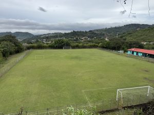 Panoramic view of a large outdoor soccer field surrounded by tall trees and distant mountains under an overcast sky.