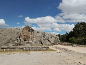 Frontal view of a large stone monolith at Qenqo, an Inca archaeological site near Cusco. The image also shows part of the surrounding area, including the stone foundations of a former Inca building built with carefully fitted blocks.