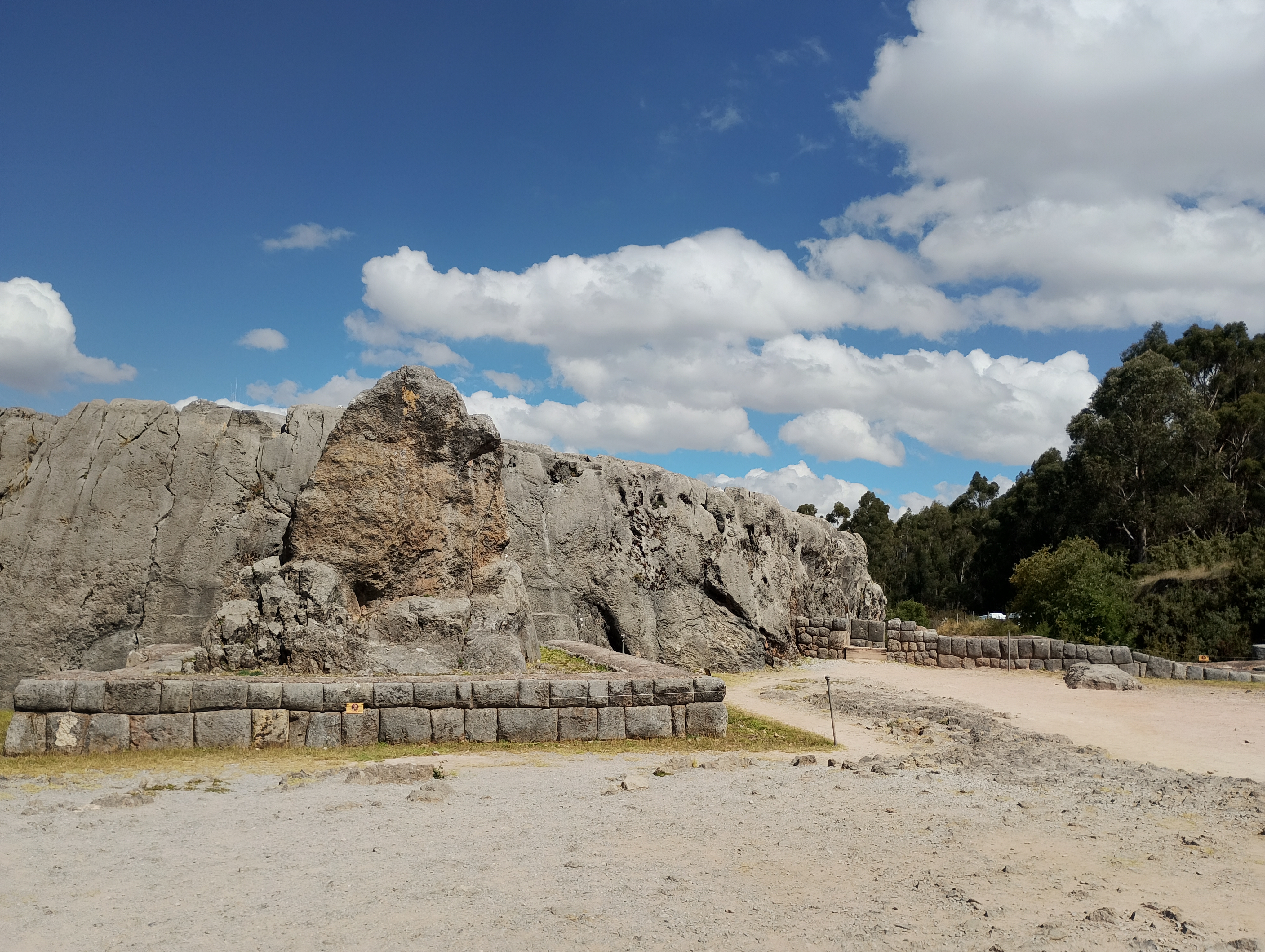 Frontal view of a large stone monolith at Qenqo, an Inca archaeological site near Cusco. The image also shows part of the surrounding area, including the stone foundations of a former Inca building built with carefully fitted blocks.