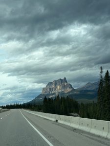 A highway through a forest leading toward a rocky mountain under storm clouds.