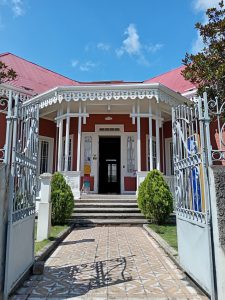 Garden entrance to a historic red Victorian-style building in Cartago, Costa Rica, now home to a UNED university center. Ornate white wrought-iron gate opens onto a tiled pathway leading to the covered entrance porch.