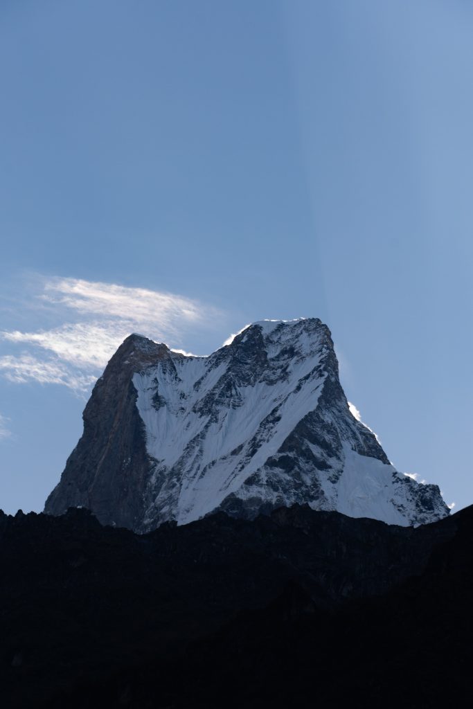 Twin-peaked – Machhapuchhre peak, snow-covered mountain rising above a dark silhouetted ridge against a blue sk