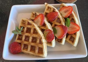 A white square dish holds four golden-brown waffles cut into triangular pieces. Each waffle is topped with bright red strawberry slices and garnished with a small mint leaf. A couple of whole strawberries sit alongside the waffles, adding a pop of color. 
