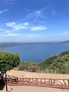 Panoramic view of a blue lake surrounded by green hills, seen from an elevated viewpoint with a winding path, decorative wooden railing, colorful posts, and flowering plants in the foreground.