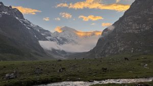 A tranquil mountain valley at sunset with a flowing stream in the foreground. Snow-capped peaks and golden clouds create a serene, majestic atmosphere.