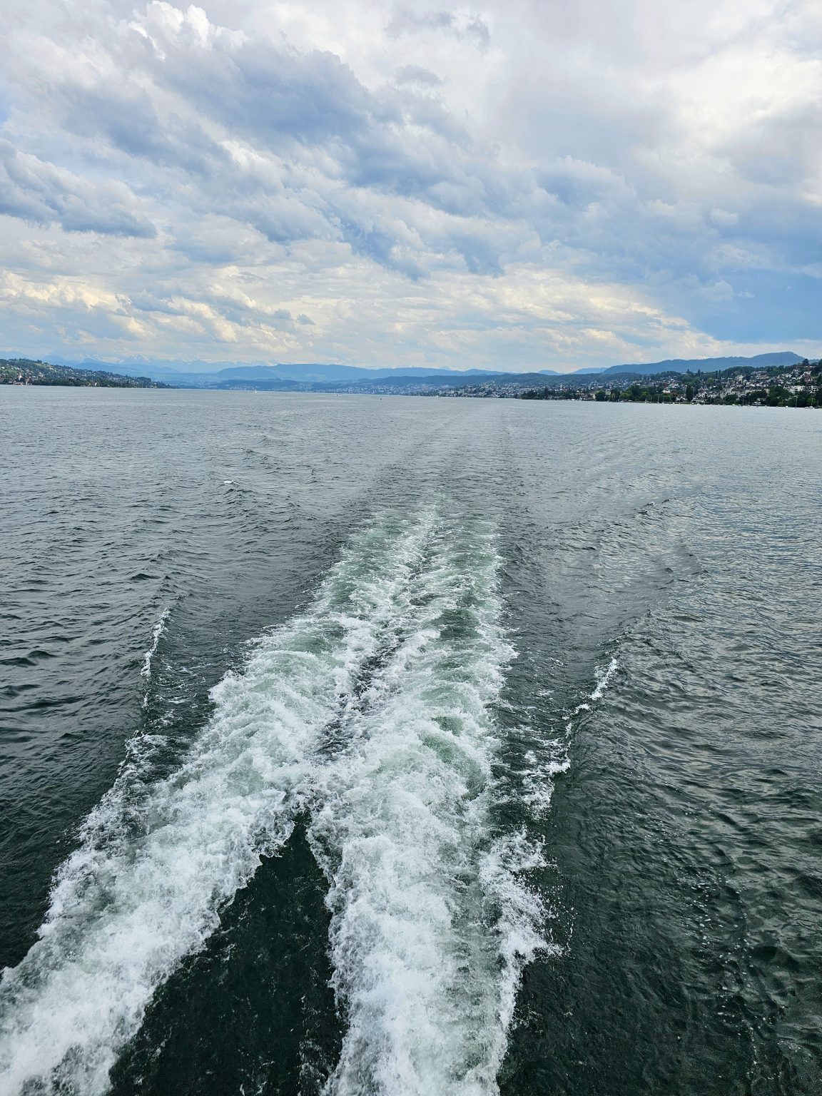 The foamy wake of a moving boat stretches across the calm waters of Lake Zurich, Switzerland, leading the eye toward distant hills beneath dramatic clouds. The peaceful landscape captures the beauty of a journey on the lake.