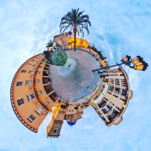 Elche at sunset: a 360° spherical photo of the Plaza de la Glorieta. In the center, the turquoise mosaic sphere and the large date palm. All around, historic terracotta buildings, the blue dome of the Basilica of Santa María, classic streetlights glowing, and passers by all set against a cloudy sky.
