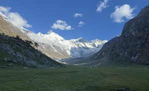 Snow-capped mountains under a clear blue sky with scattered clouds tower over a lush green valley. The serene landscape conveys tranquility.