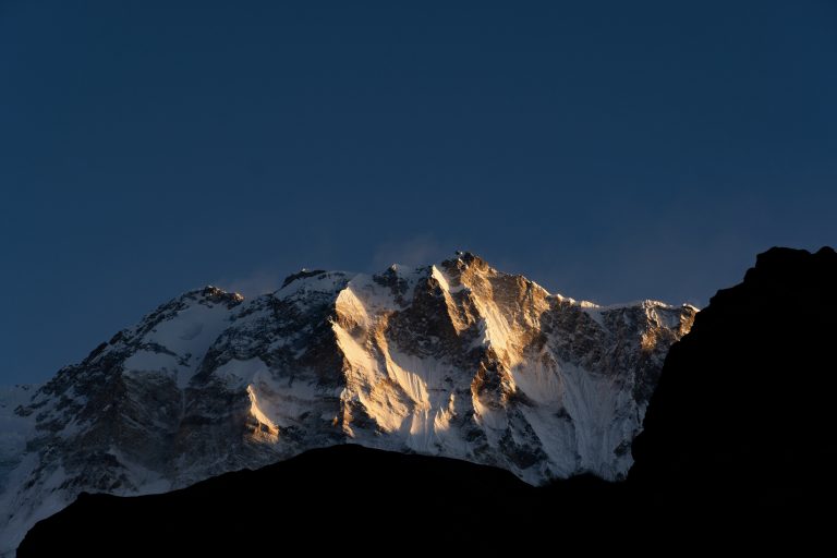 Snow-capped mountain peak bathed in golden sunrise light against a deep blue sky.