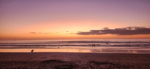Peaceful beach sunset with a person on the sand, people in the water, and colorful evening sky.