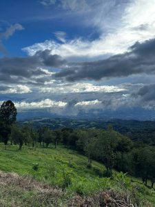 A wide view of green hills and trees overlooking a valley, with low clouds and dramatic blue-and-gray skies above