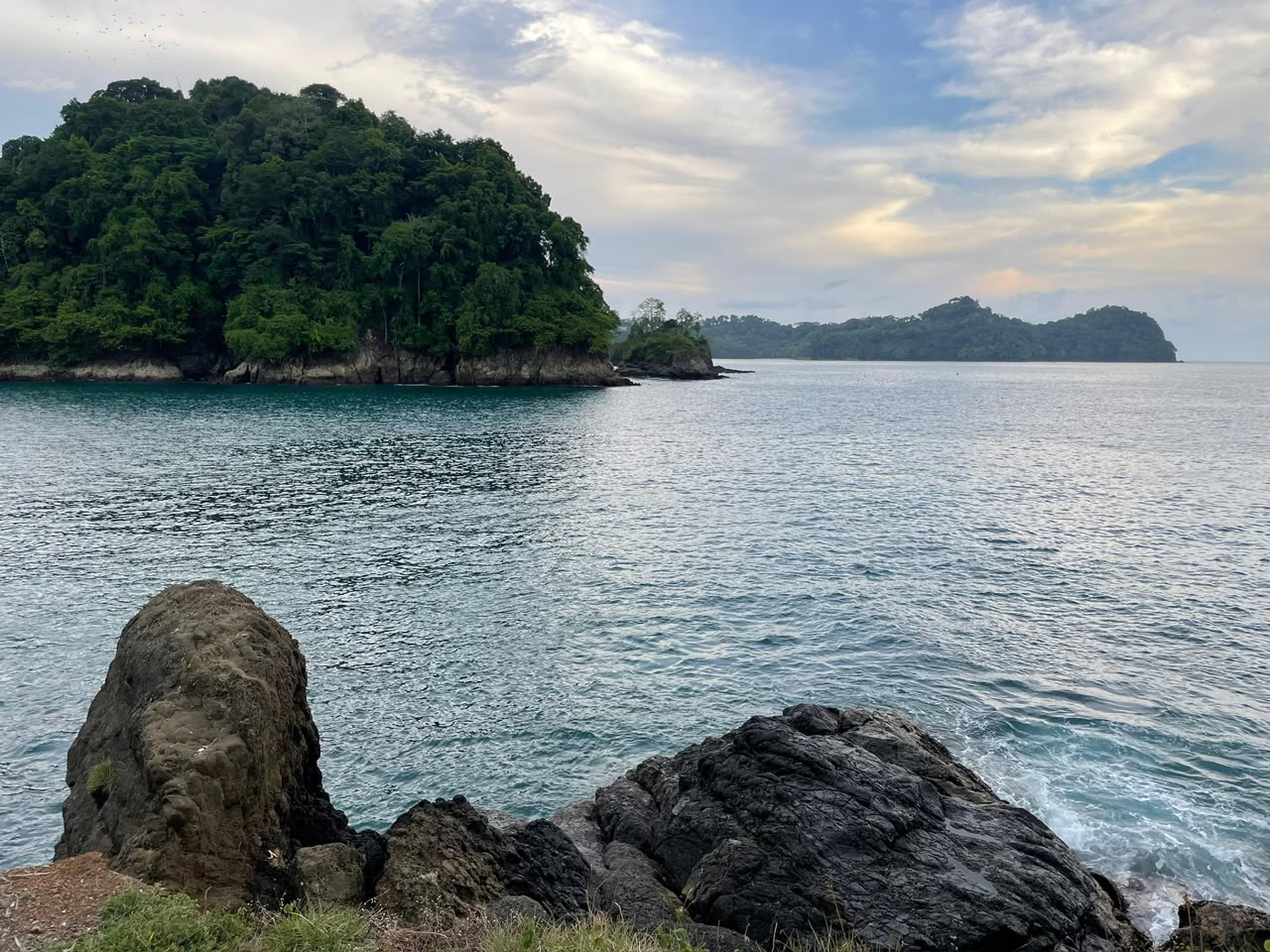 Calm coastal water with rocky shore, a green island, and a partly cloudy blue sky.