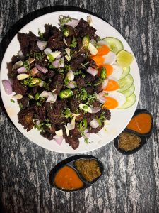 White plate of crispy dark meat known as sekuwa, garnished with onions, green chilies, and cilantro, served with carrot, cucumber, and lime slices, alongside two dipping sauces on a stone-textured surface.