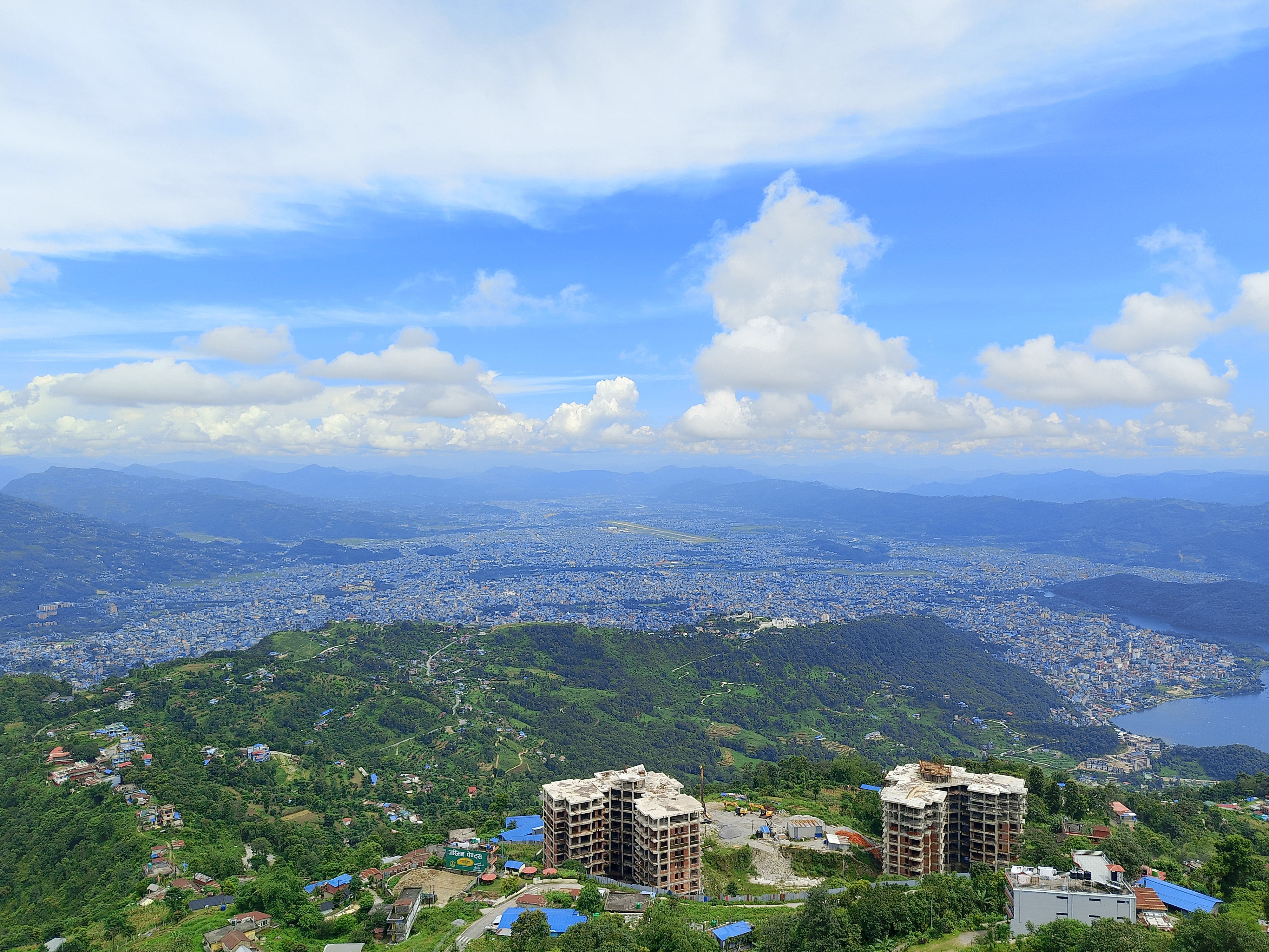 Aerial view of Pokhara city and airport runway nestled in a valley, surrounded by green hills and distant mountains under a partly cloudy sky