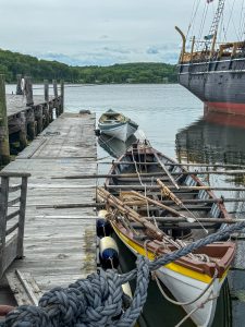 Two old row boats with oars and whaling harpoons lined up against the pier at the Mystic Seaport Museum. Part of a tall ship is off to the right.
