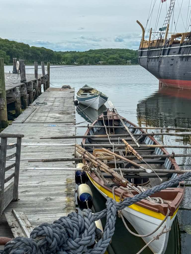 Two old row boats with oars and whaling harpoons lined up against the pier at the Mystic Seaport Museum. Part of a tall ship is off to the right.