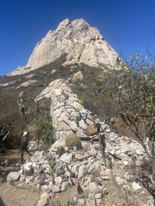 Stone structure in the foreground with Peña de Bernal rising in the background under a clear blue sky in Querétaro, Mexico.