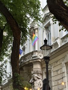 Rainbow flag displayed on the balcony of a historic building framed by trees and a street lamp in Bratislava, Slovakia.