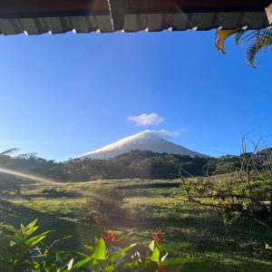 A scenic view framed by the edge of a building’s roof shows a majestic volcano in the distance, partially covered by wispy clouds.
