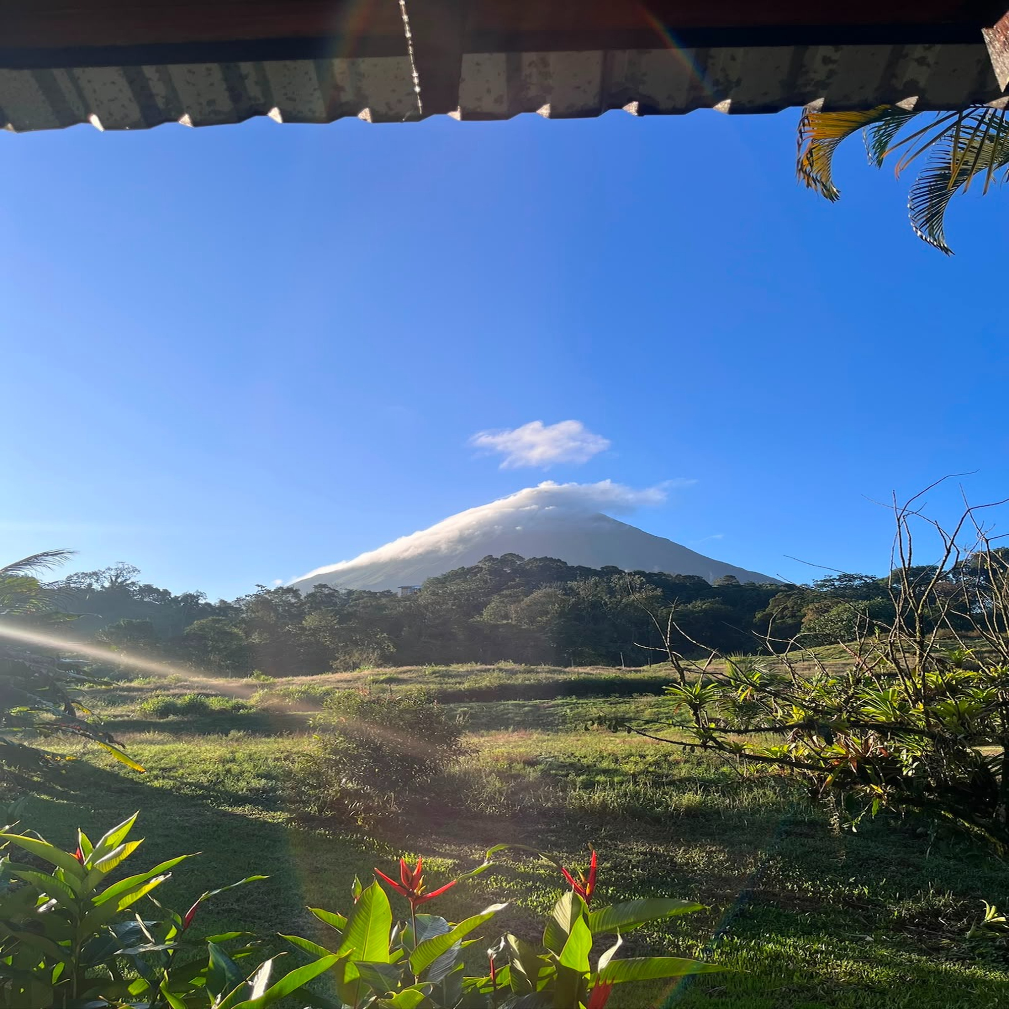 A scenic view framed by the edge of a building's roof shows a majestic volcano in the distance, partially covered by wispy clouds.