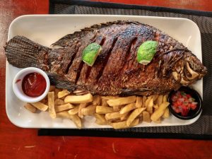 A whole grilled tilapia served with French fries, lime wedges, tomato salsa, and dipping sauce on a rectangular plate.
