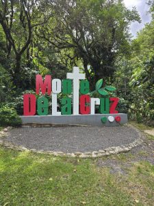Large red, green, and white 3D sign reading "Monte De La Cruz" set against a lush forest backdrop.
