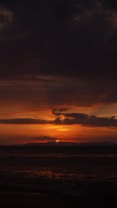 A deep orange sunset over the ocean, with dark clouds above and the sun setting behind distant mountains on the horizon.