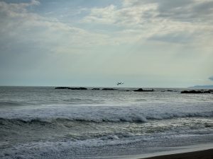 The image captures a serene coastal scene, featuring gentle waves rolling toward the shore on a sandy beach. In the distance, a group of dark rocks emerges from the water, contrasting with the lighter hues of the ocean. Jaco Beach, Costa Rica