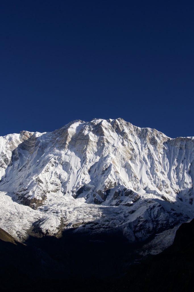 Wide snow-covered mountain massif with vertical rock ridges under a dark blue sky, dark foreground silhouette below