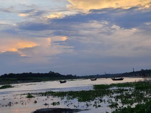 Small boats float on a tranquil river under a cloudy twilight sky, with distant treelines framing the horizon and lush green plants in the foreground.
