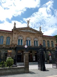 Front facade of the National Theater of Costa Rica in San José, featuring ornate stone carvings, arched windows, wrought-iron gates and rooftop statues crowning the pediment above the main entrance.
