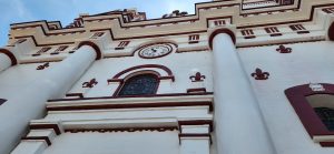Low-angle view of ornate church architecture with red accents, large columns, arched windows, and a round clock set above the entrance.