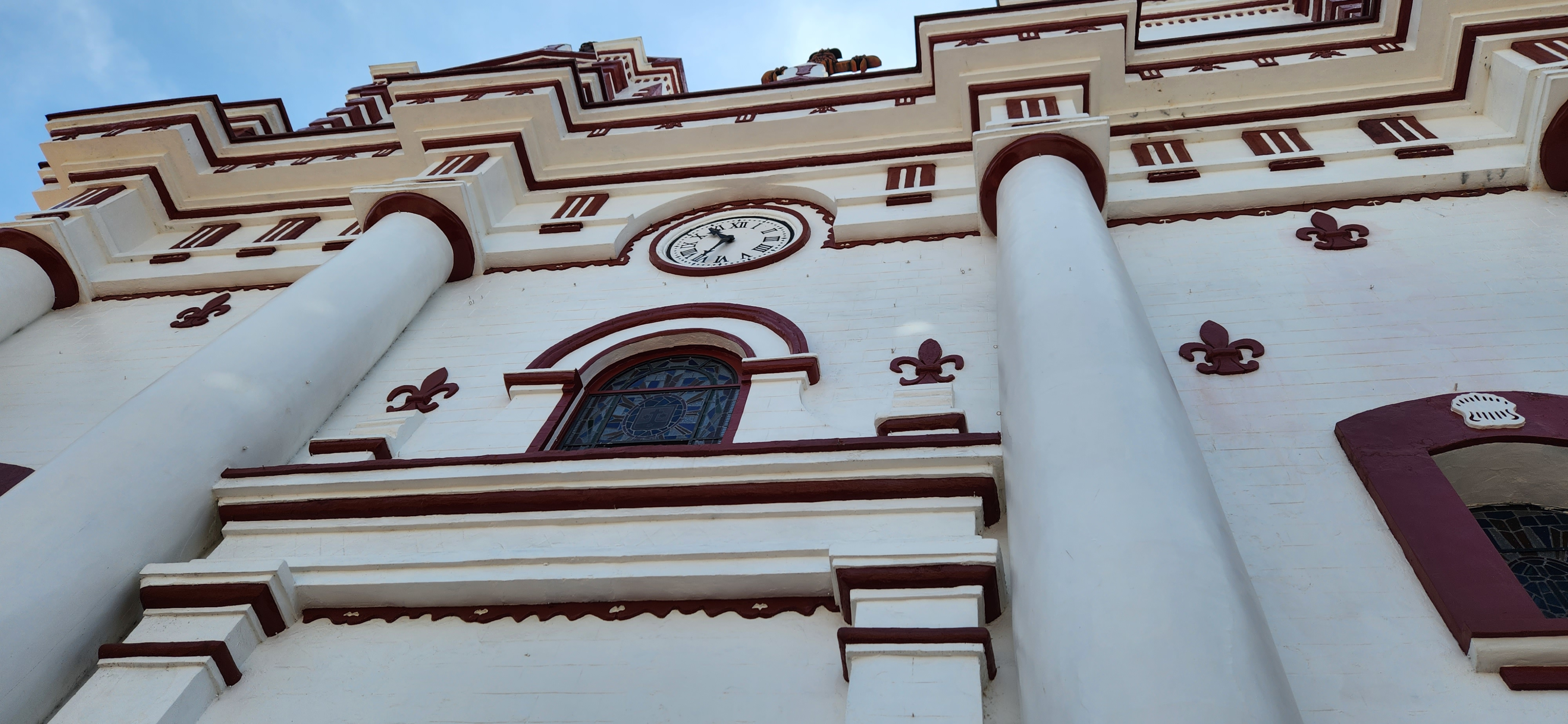 Low-angle view of ornate church architecture with red accents, large columns, arched windows, and a round clock set above the entrance.