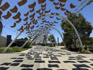 Decorative pedestrian walkway outside Optus Stadium in Perth, featuring curved white arches with artistic shade panels casting patterned shadows across the paved pathway on a sunny day.
