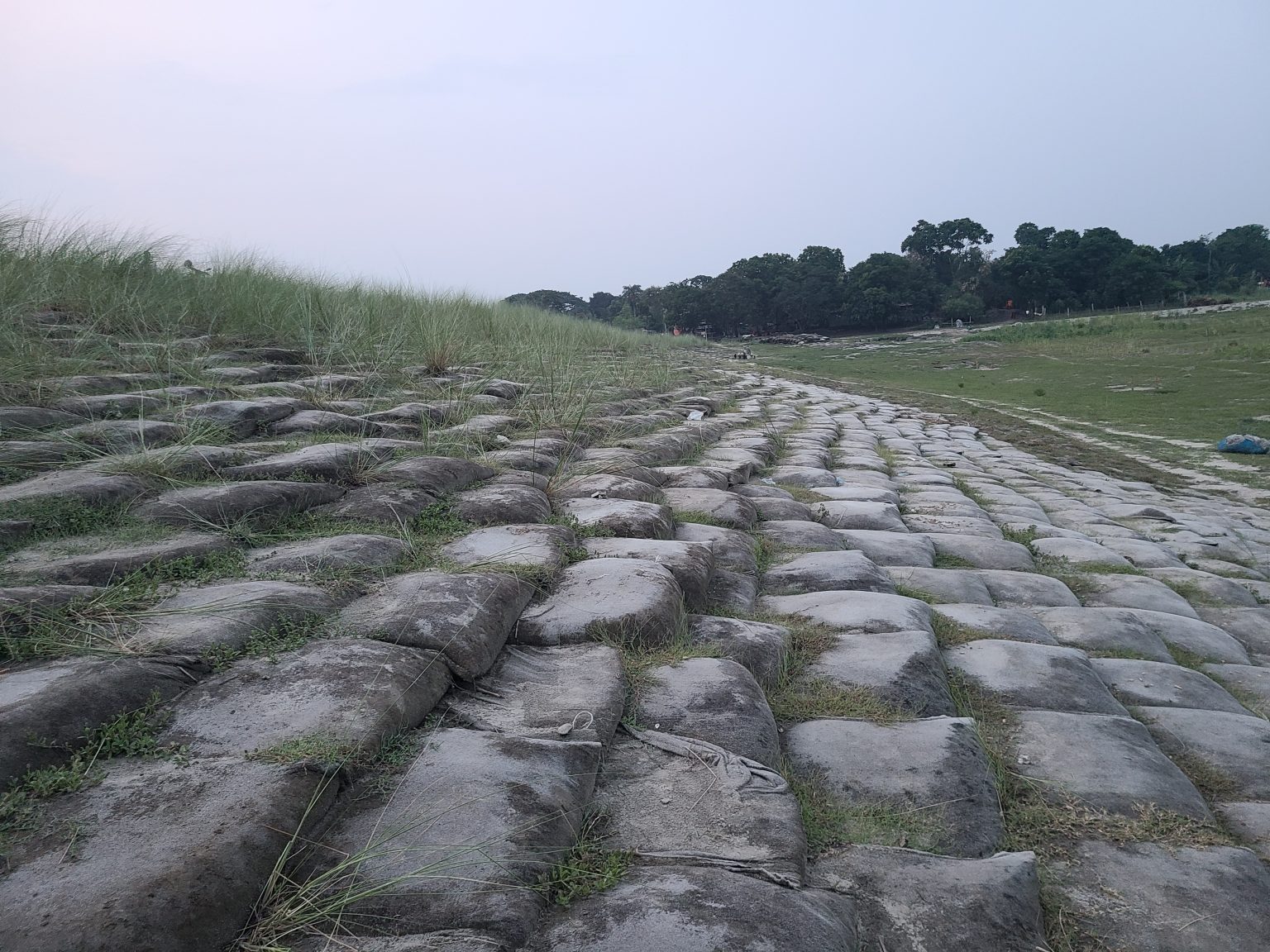 A low-angle, wide shot shows a grassy, stepped bank lined with sandbags, leading down to a dry riverbed under an overcast sky.