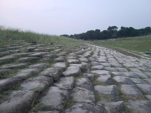A low-angle, wide shot shows a grassy, stepped bank lined with sandbags, leading down to a dry riverbed under an overcast sky.
