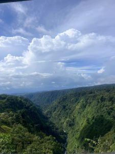 A wide green valley stretches between forested hills under a bright sky filled with large white clouds.