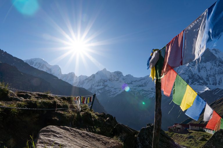 Colorful prayer flags strung on a rocky ridge with snow-capped peaks and small buildings below, sun shining brightly in the sky