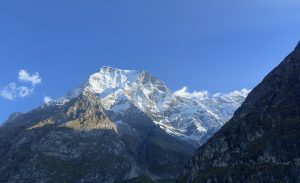 Snow-capped mountain peak under a clear blue sky, flanked by rugged cliffs. Soft sunlight highlights the snowy textures, evoking a serene, majestic feeling.