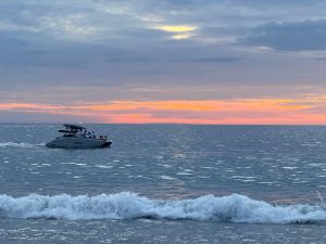 A sleek, white motorboat is cruising across a calm sea, carrying several people. The water glistens under the soft glow of a colorful sunset, with hues of pink, orange, and purple blending in the sky. Gentle waves are rolling towards the shore, creating a peaceful atmosphere. In the distance, a small silhouette of a boat can be seen, far away on th
