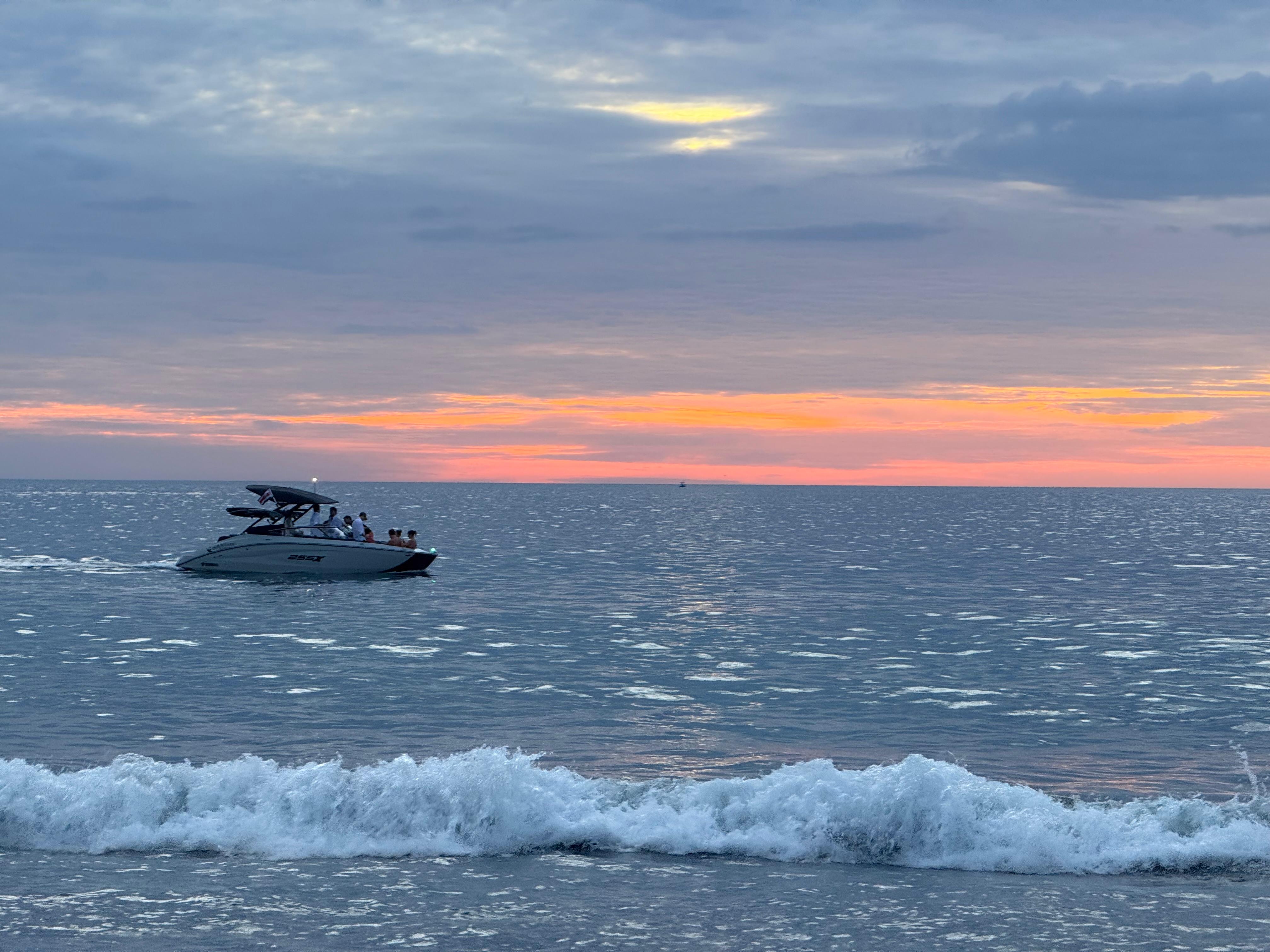 A sleek, white motorboat is cruising across a calm sea, carrying several people. The water glistens under the soft glow of a colorful sunset, with hues of pink, orange, and purple blending in the sky. Gentle waves are rolling towards the shore, creating a peaceful atmosphere. In the distance, a small silhouette of a boat can be seen, far away on th