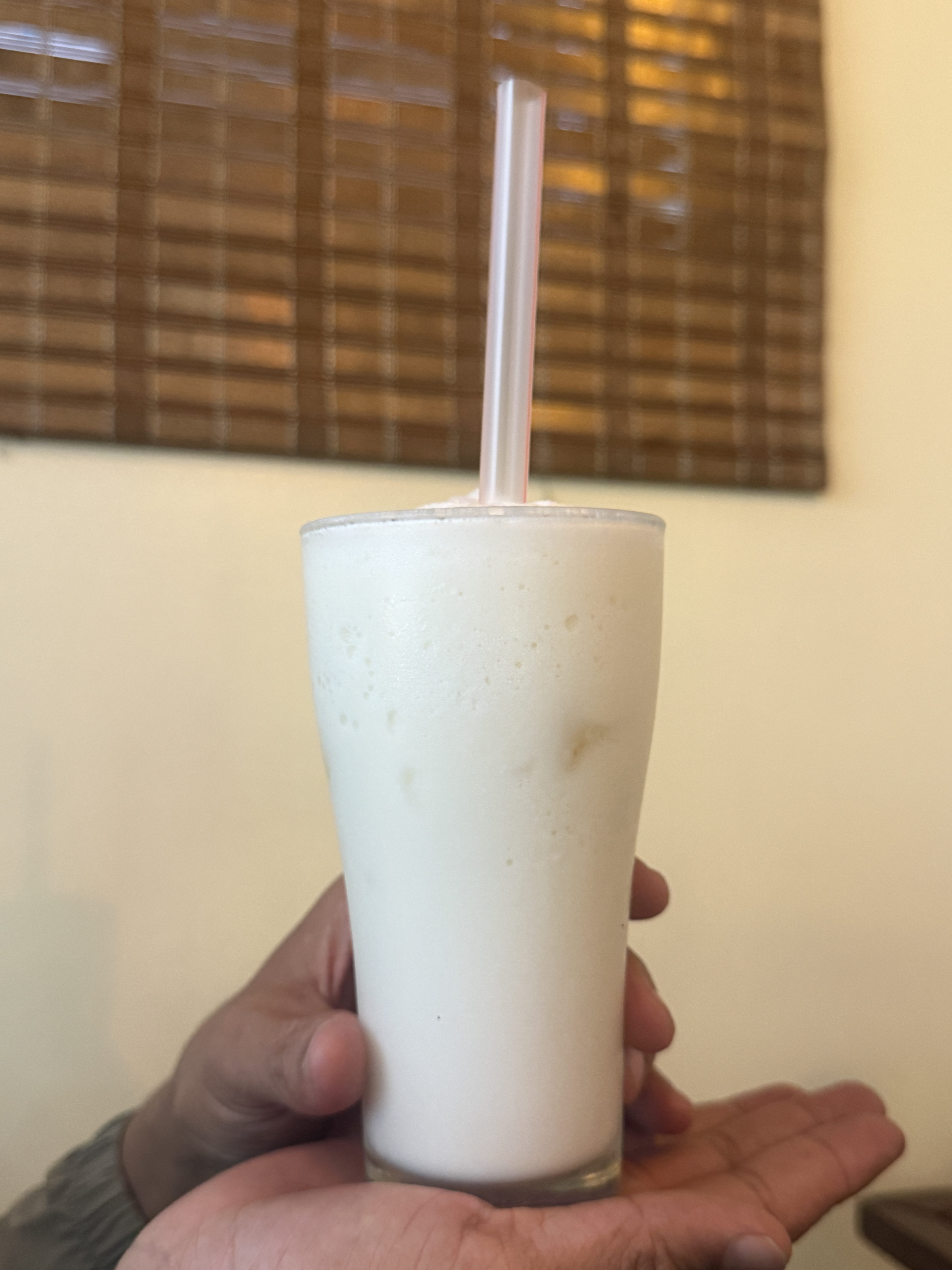 A hand holding a glass of creamy white beverage with a straw, set against a softly lit interior with a wooden blinds background.