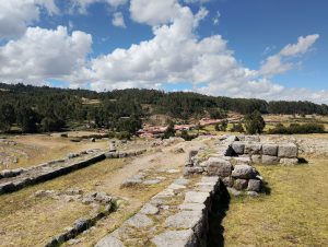 Stone foundations of an ancient Inca building at Sacsayhuaman, with small buildings and a forest in the distance.